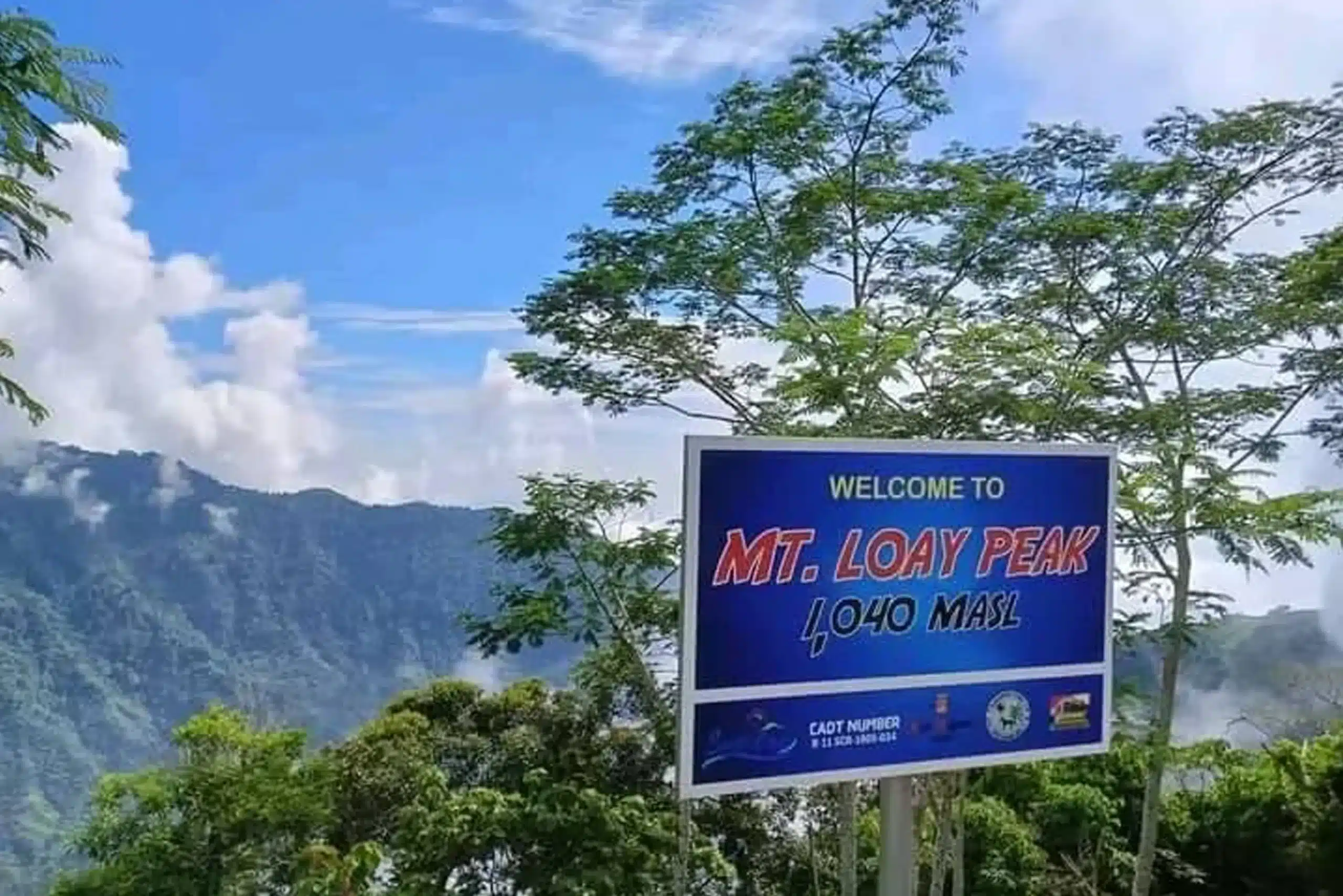 A blue welcome sign for Mt. Loay Peak in Sta. Cruz, Davao del Sur, showing an elevation of 1,040 MASL against a backdrop of lush green mountains and a cloudy blue sky.