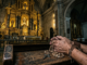 Close-up of calloused, weathered hands holding a wooden rosary on a worn pew in a dimly lit Philippine cathedral, with a blurred opulent gold altar in the background
