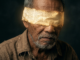 A hyper-realistic close-up portrait of an elderly Filipino man with a pensive expression, wearing a worn linen shirt. A blindfold made of aged, glowing parchment with Latin scripture covers his eyes, set against a dark, moody background with dramatic cinematic lighting.