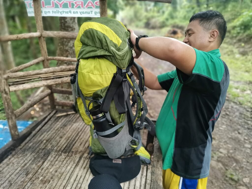 A focused close-up photograph of a smiling Filipino hiker (a 'Manakaay' explorer) adjusting the straps of his substantial green and yellow overnight trekking backpack at a rustic wooden shelter near the Mt. Dinor jump-off point in Brgy. Sinoron, Sta. Cruz. The image highlights the detailed gear preparation required for challenging climbs in Davao del Sur